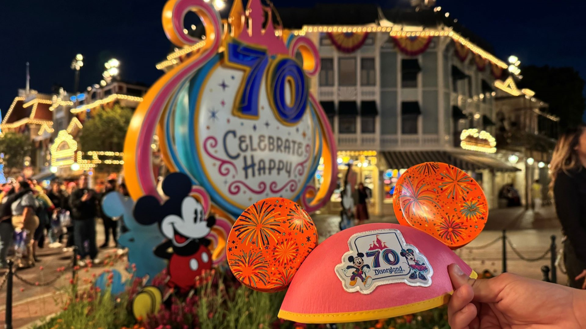A Disneyland 70 Mickey Mouse ear hat is held up in front of a larger sign for Disneyland 70 at Disneyland Park