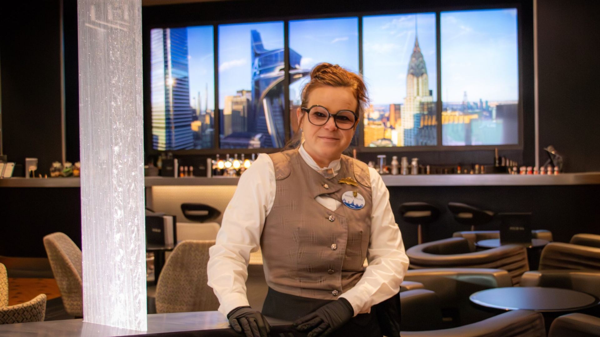 A Disney cast member in glasses looks at the camera and behind her is an image of Avengers Tower