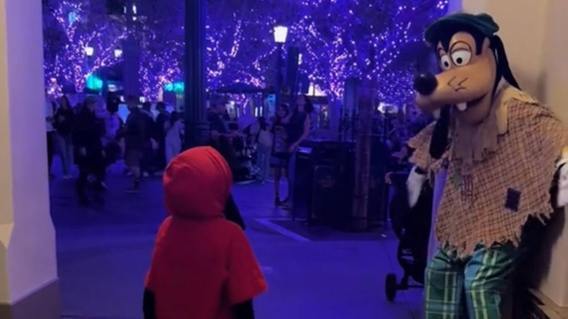 Goofy looks at a child in front of lit-up purple trees.