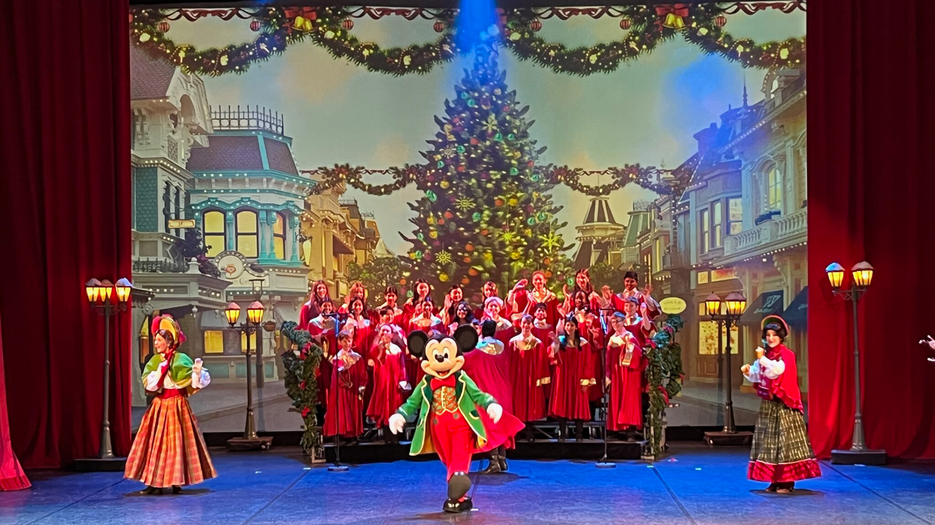 Mickey Mouse stands in front of a choir with the image of a Christmas tree on Main Street U.S.A. as the backdrop