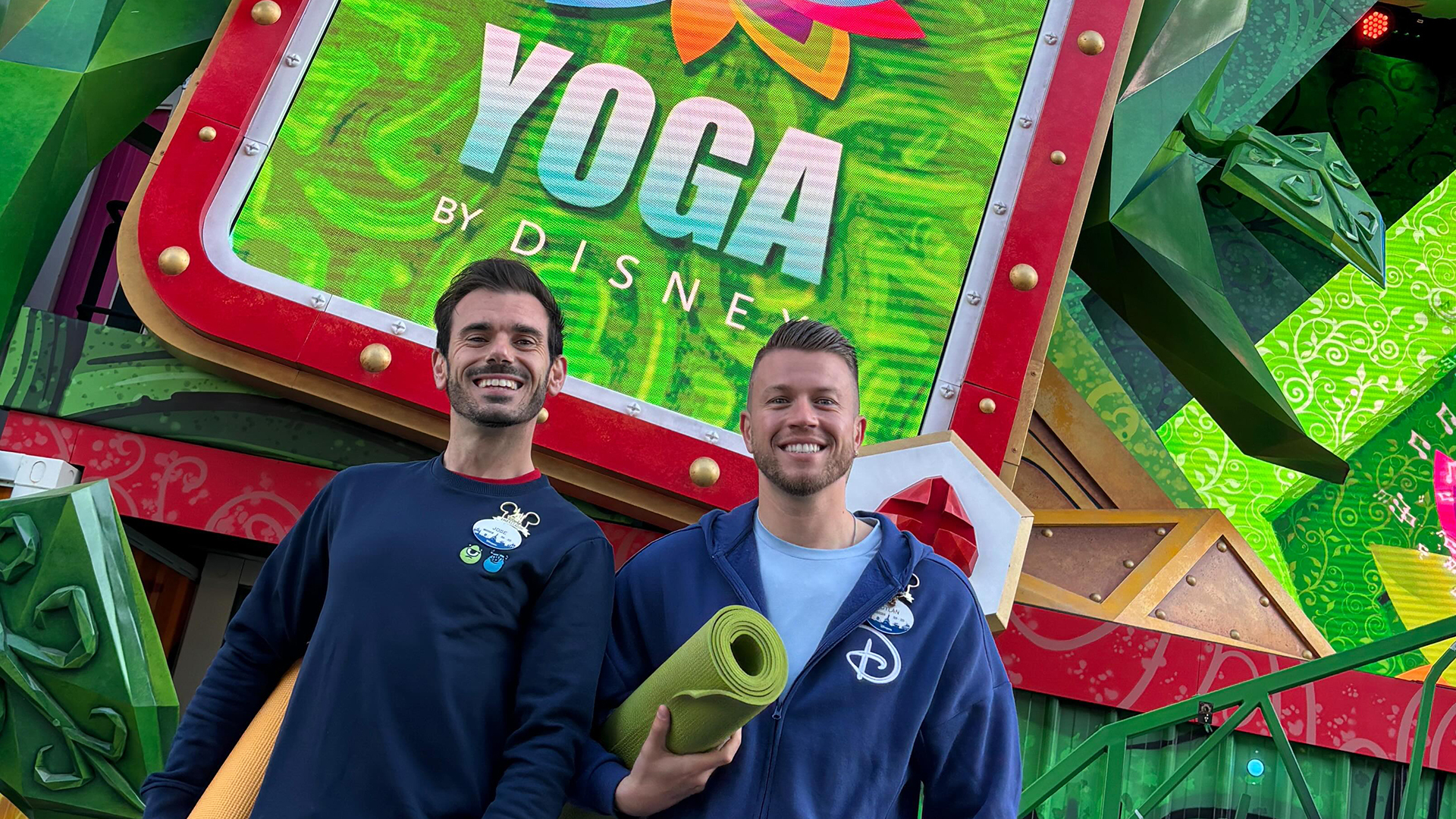 Two men stand in front of a Yoga by Disney sign