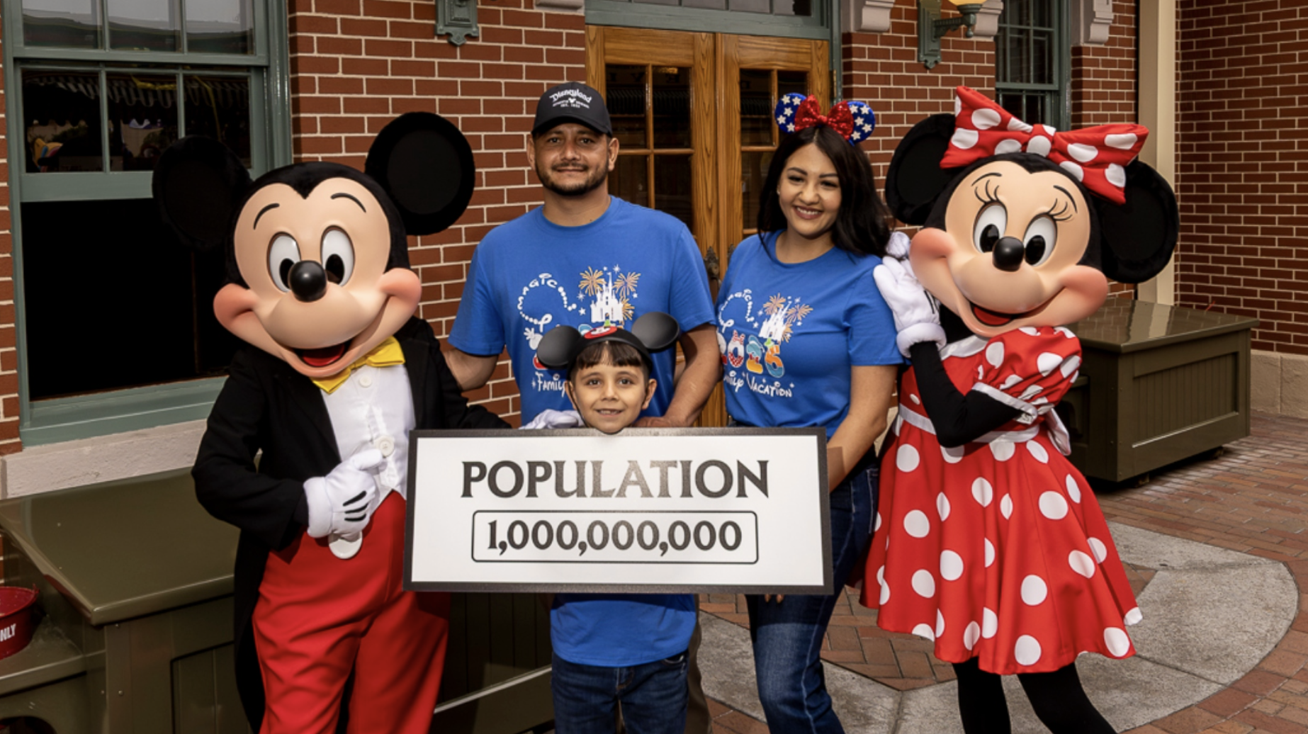 Mickey Mouse and Minnie Mouse stand with a family of three holding a sign that reads &ldquo;Population 1,000,000,000&rdquo; in front of a brick building at Disneyland Resort, celebrating the resort&rsquo;s one billionth guest milestone.