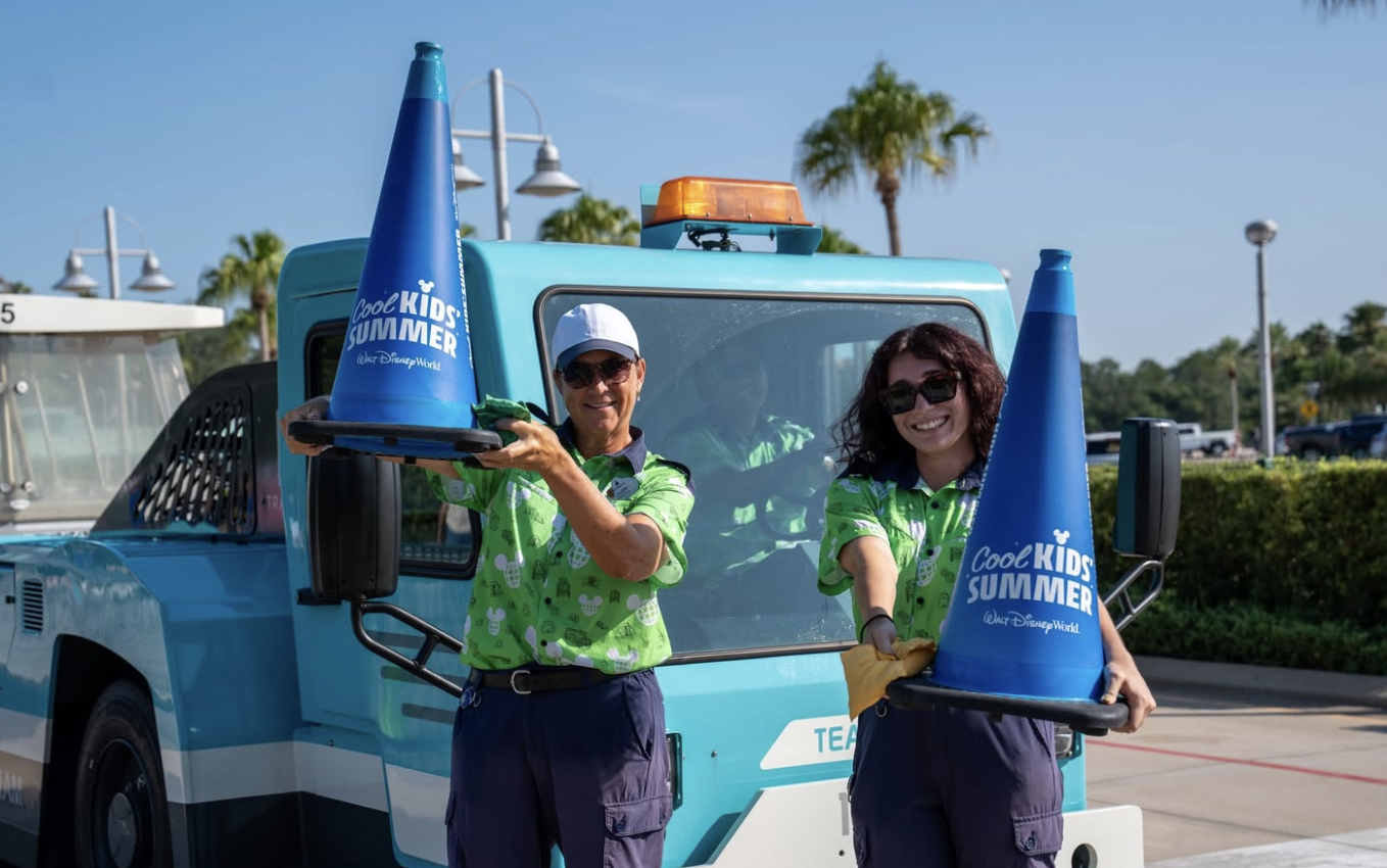 Two Walt Disney World Cast Members stand in front of a blue service vehicle holding oversized blue traffic cones branded with the &ldquo;Cool Kid Summer&rdquo; Walt Disney World logo. Palm trees and a sunny outdoor setting are visible in the background.