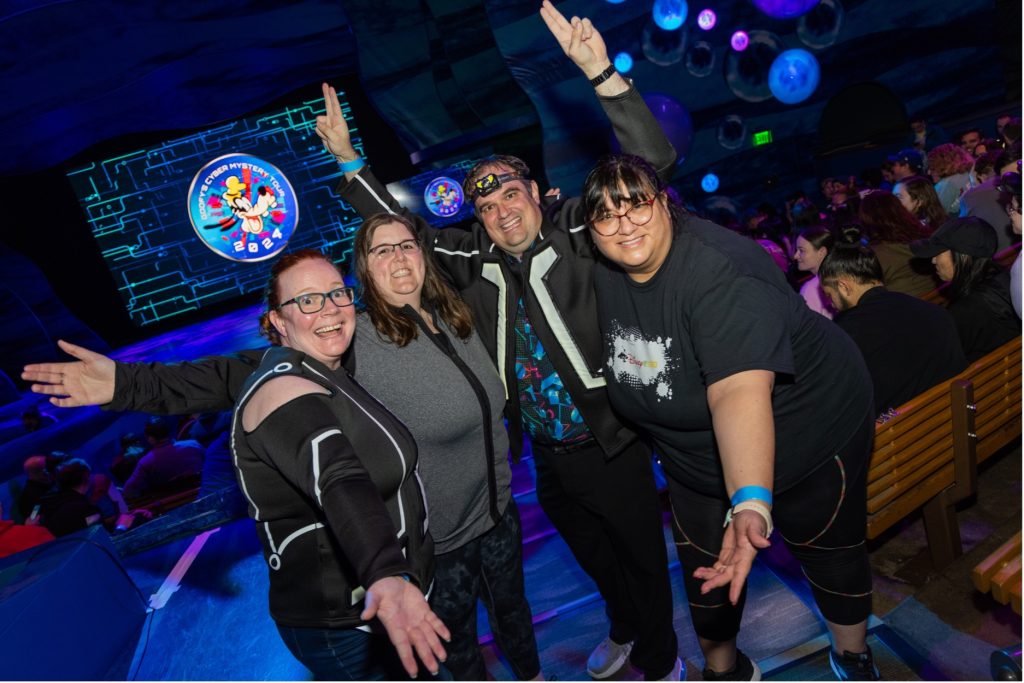 Four cast members standing in front of a goofy's mystery tour sign