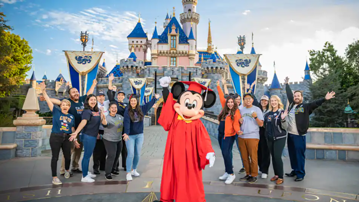 Mickey wearing red graduation robes standing outside Sleeping Beauty Castle with a dozen Aspire students.