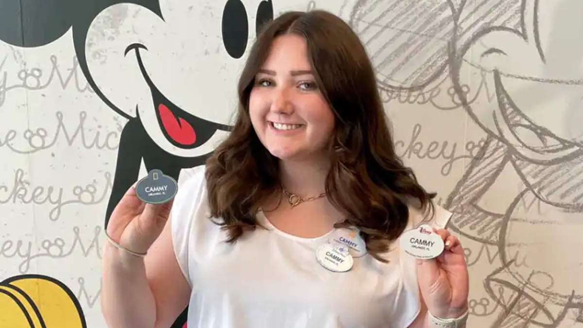 Woman with brown hair, wearing a white shirt is holding up her 4 Disney nametags in front of a Mickey sketch background.